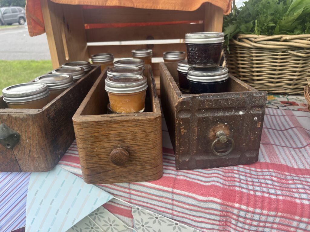 Three card file drawers filled with small mason jars of orange pumpkin butter and amber maple syrup on a table with a pink checked cloth from a farmer's market.