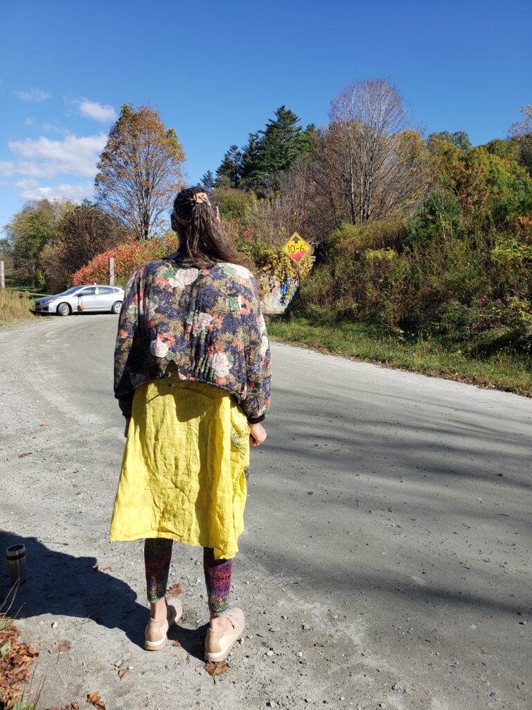 A woman stands with her back to the camera. She wears a yellow dress and a flower print jumper. She is looking over a fall display of final color, waiting for the train to pass.