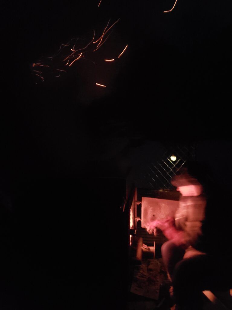A person sits in front of a fire, they are glowing red and sparks are flying above their head. The image is slightly blurry giving it an otherworldly feel